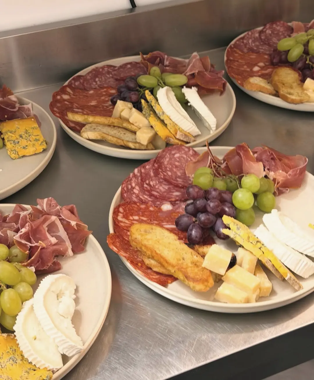 Plates of salami, prosciutto, blue cheese, soft white cheese, hard cheese, grapes and toasted bread lined up on a steel counter