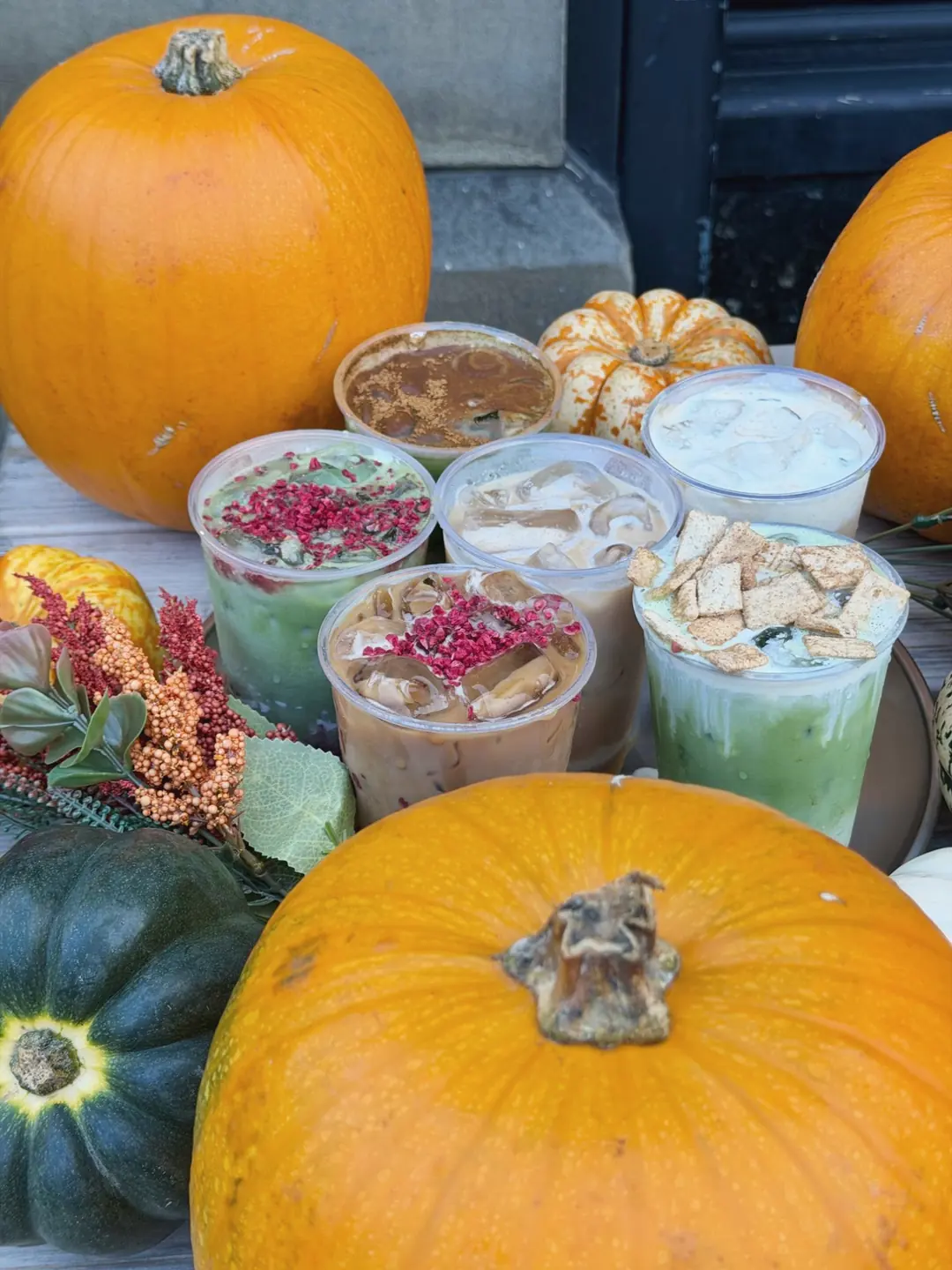 Iced matchas and lattes topped with raspberry crumb and cinnamon cereal, with a big pumpkin in front and autumn flowers beside them