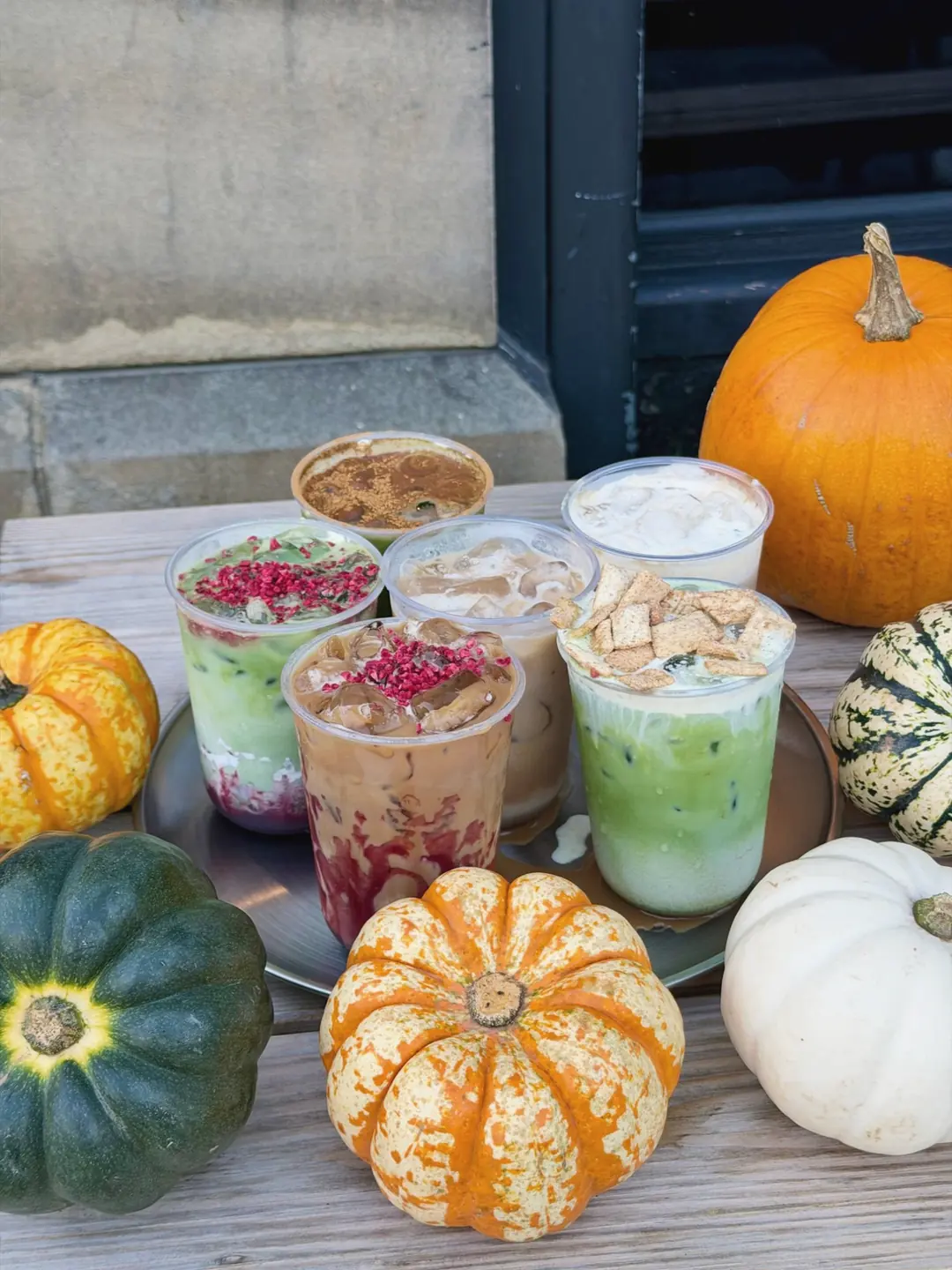 Six iced lattes and matchas on a steel tray on a wooden table, among orange, green and white pumpkins