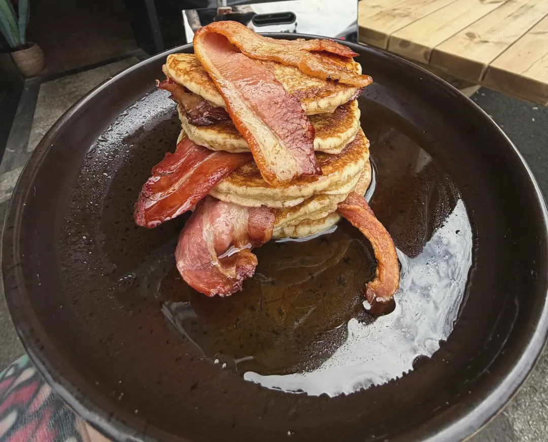 A stack of pancakes with streaky bacon and maple syrup on a dark plate, outside the café
