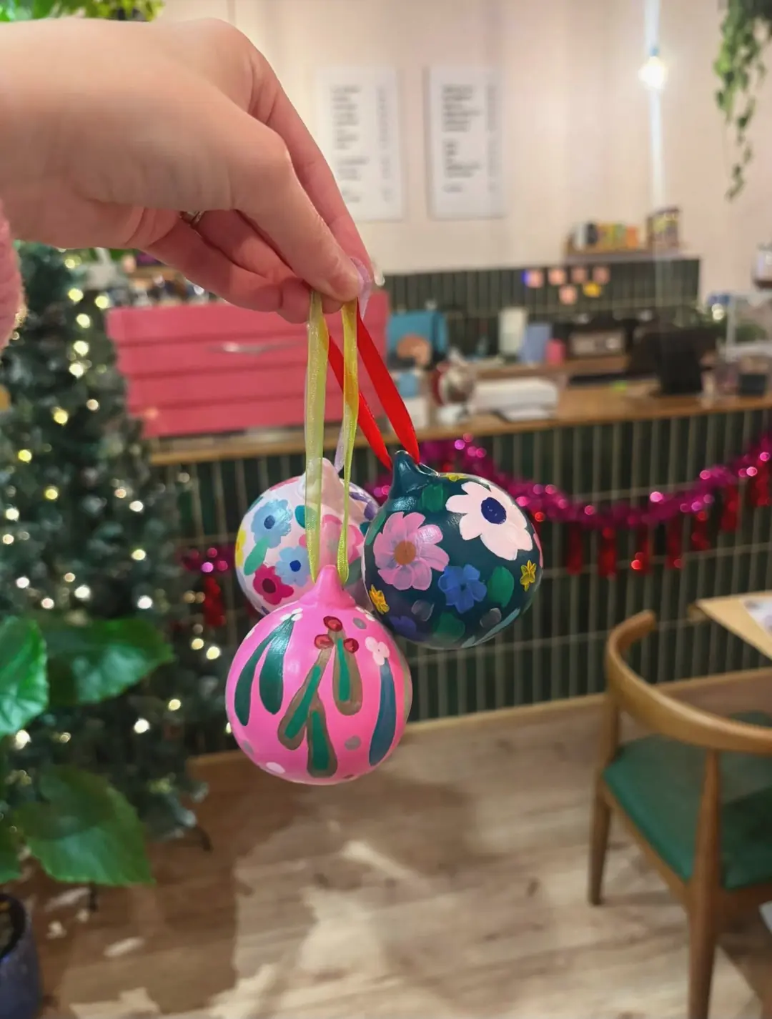 Three hand-painted ceramic baubles on ribbons, in pink, white and dark green with flowers and mistletoe, held up in front of a lit Christmas tree