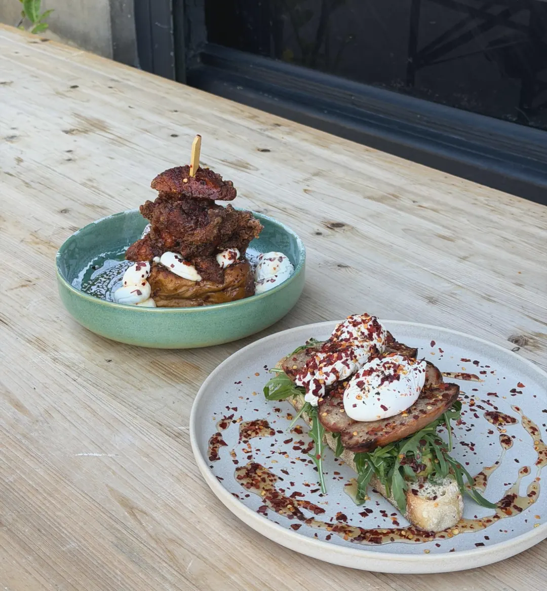 Fried chicken on French toast in a green bowl, and poached eggs and sausage on sourdough with rocket and chilli flakes, on a table outside