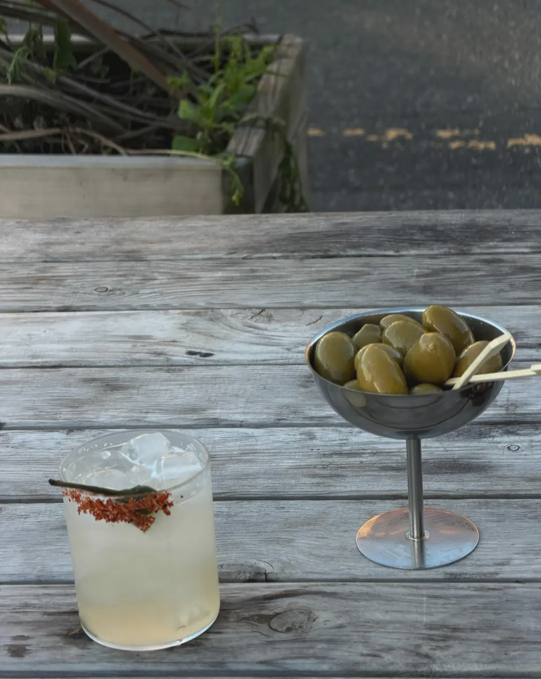 A spicy margarita with a chilli-salt rim and a green chilli, next to a bowl of olives on an outdoor table