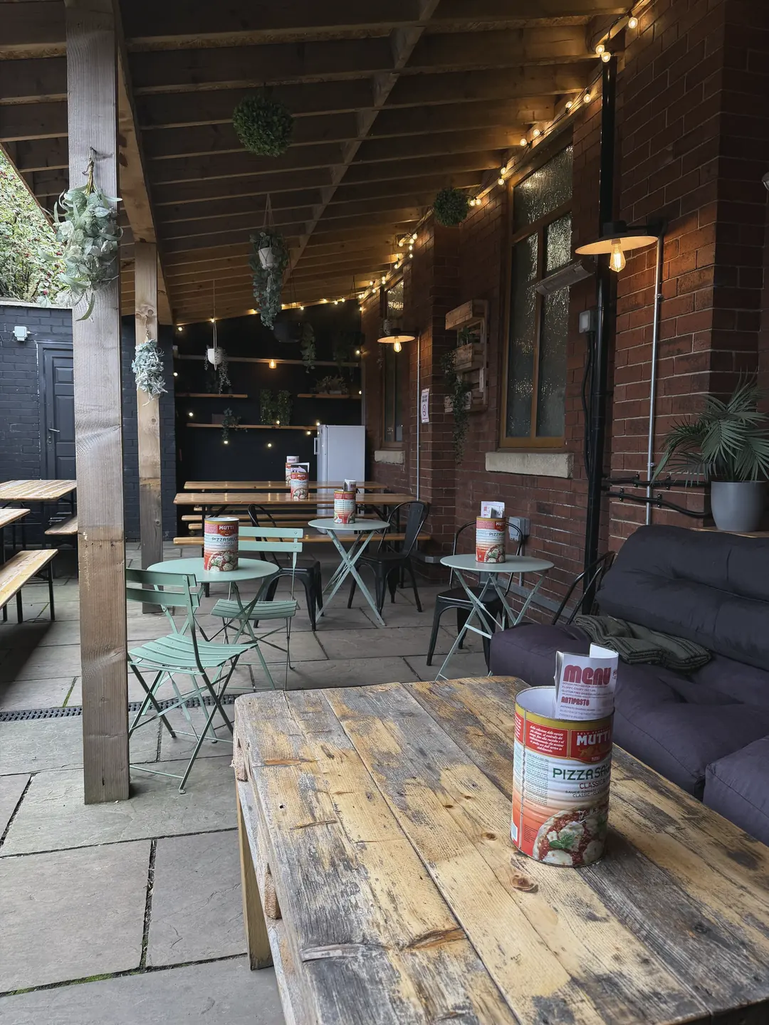 The covered courtyard at Hidden: festoon lights along the timber beams, hanging plants, bistro tables and benches, and a sofa against the brick wall