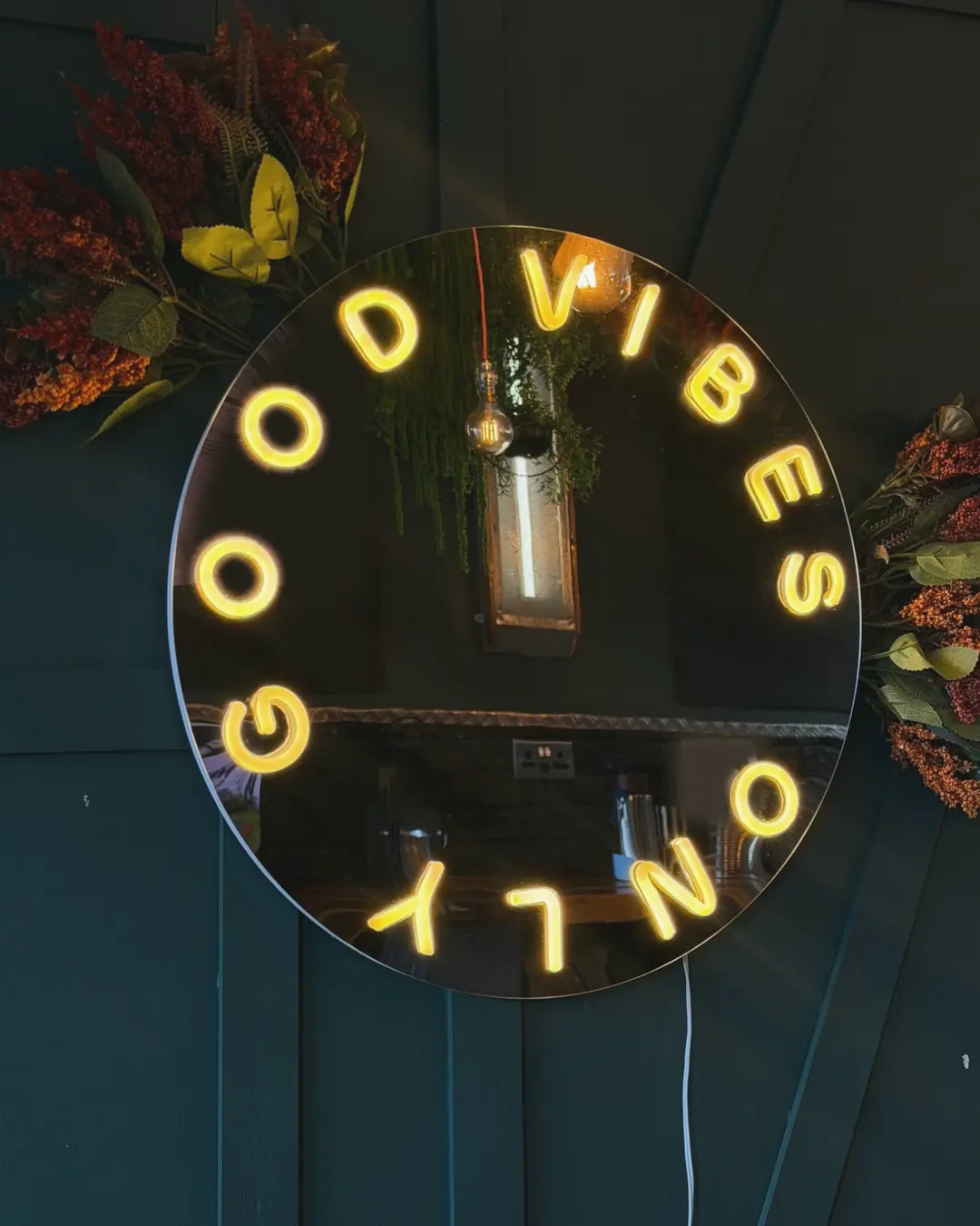 A round mirror with a warm neon sign reading Good vibes only, framed by autumn flowers on a dark green wall