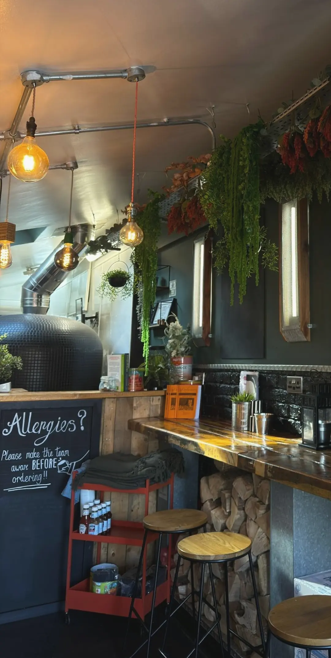 Inside Hidden: bulb lights and hanging plants over the tiled pizza oven, the Allergies chalkboard, the wooden counter and bar stools