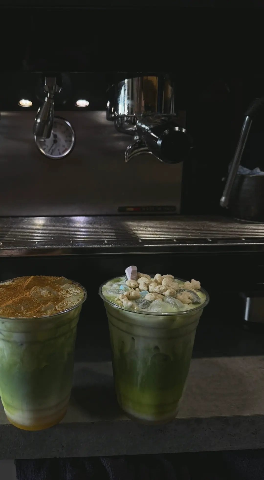 Two iced matchas, one dusted with spice and one topped with cereal, in front of the espresso machine