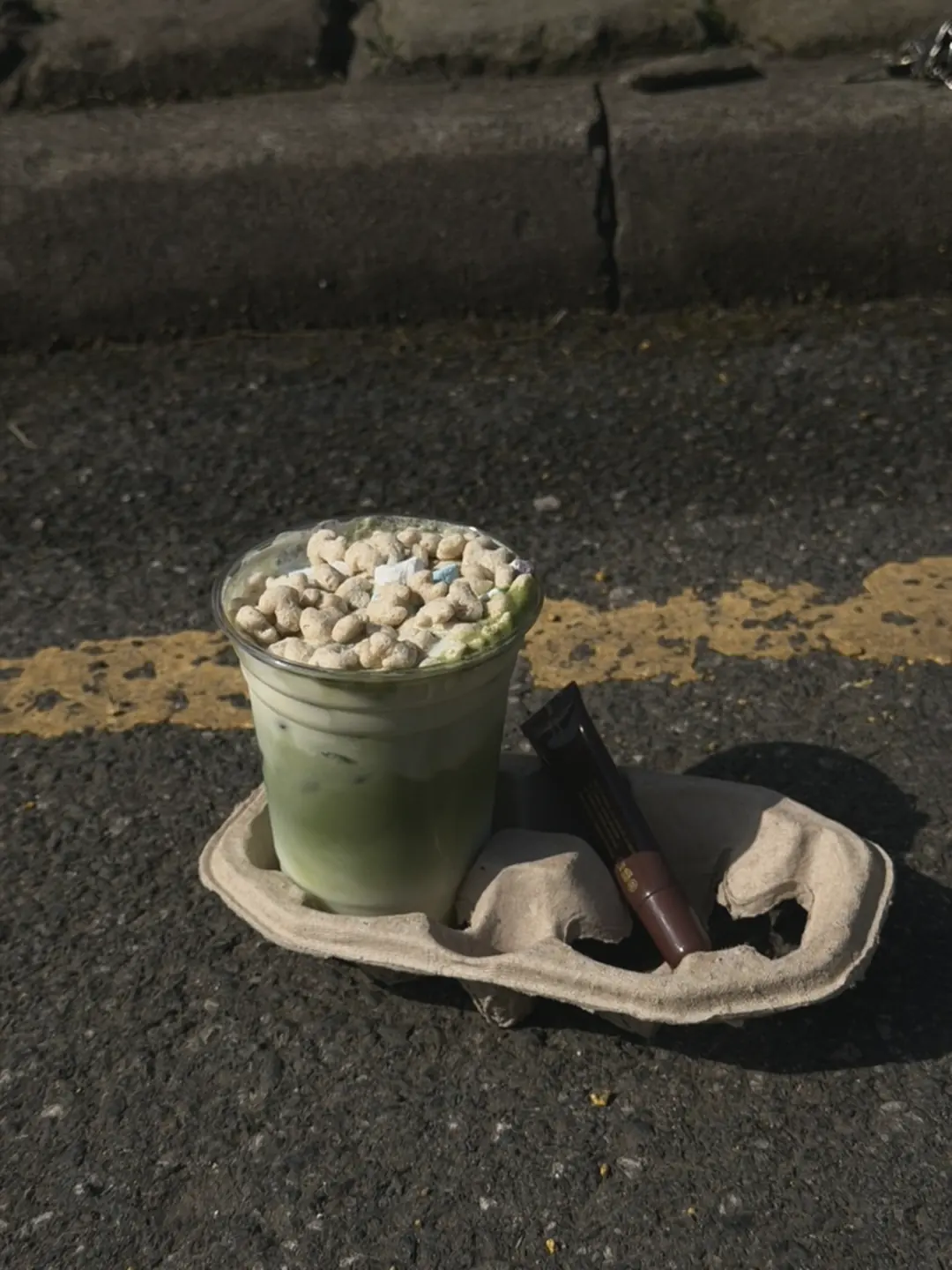 An iced matcha topped with cereal in a takeaway tray, out in the sun