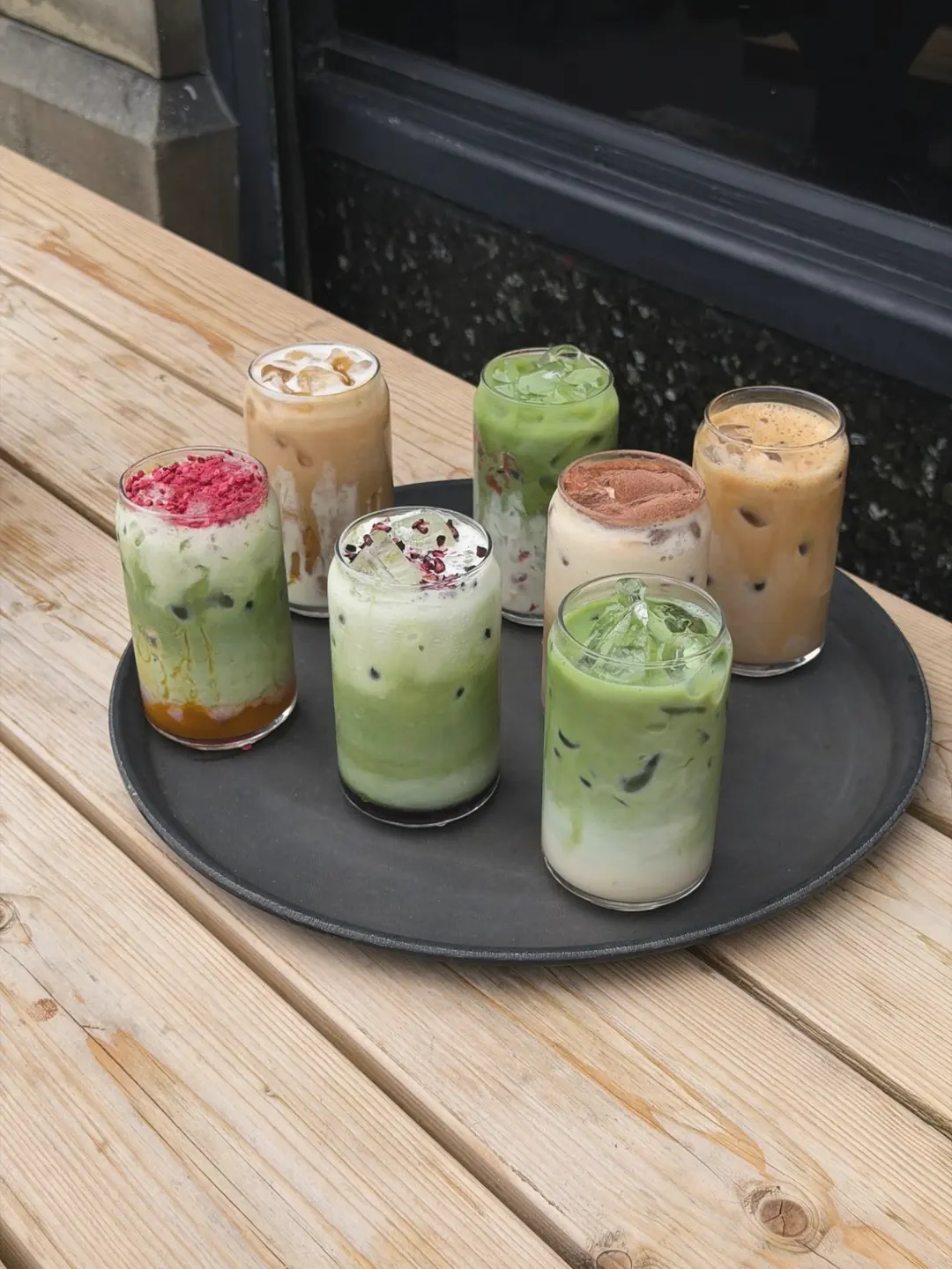 Seven iced matchas and lattes in glasses on a round tray, on a wooden table outside