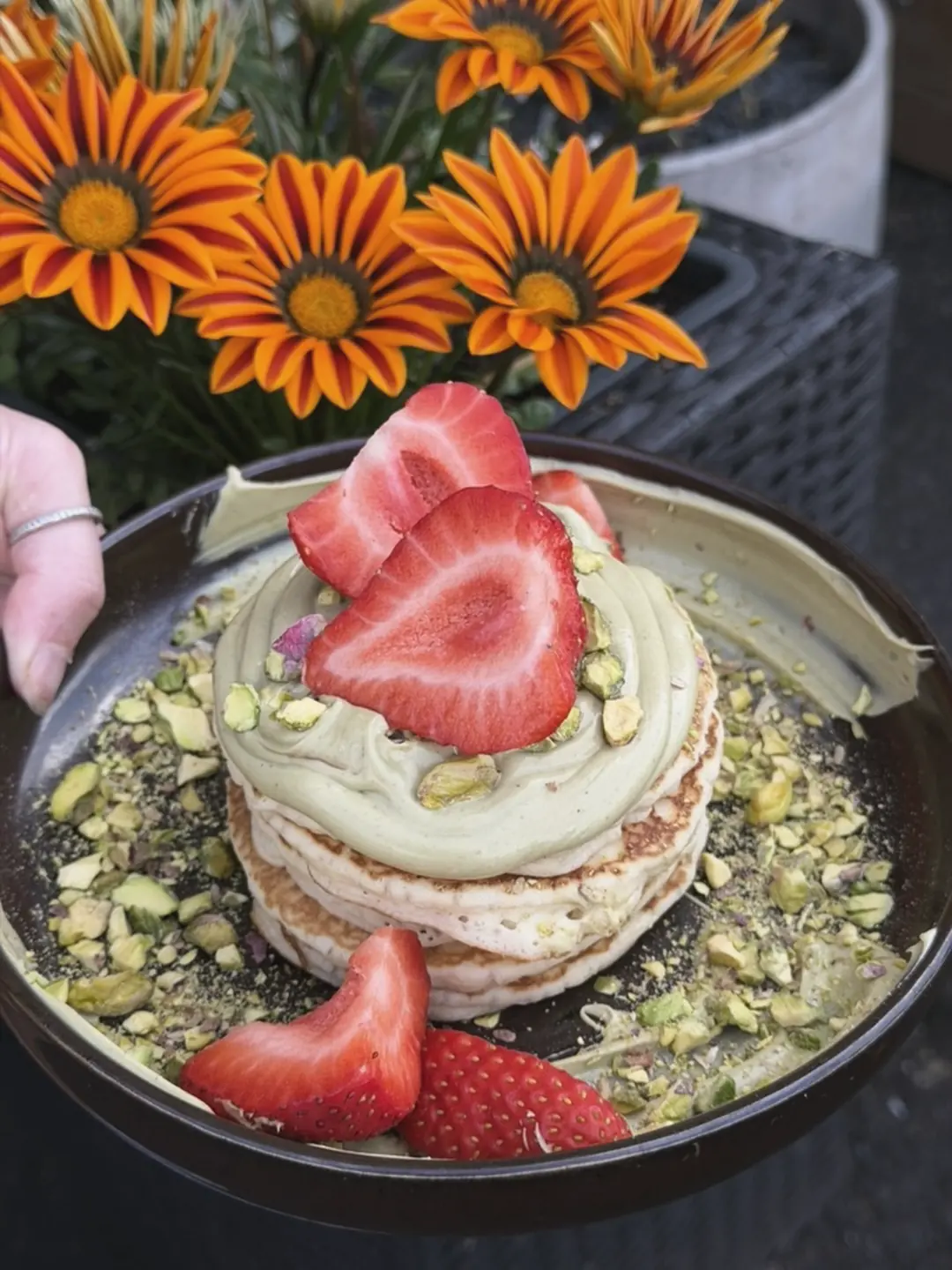 A stack of pancakes with pistachio cream, crushed pistachios and strawberries, in front of orange flowers