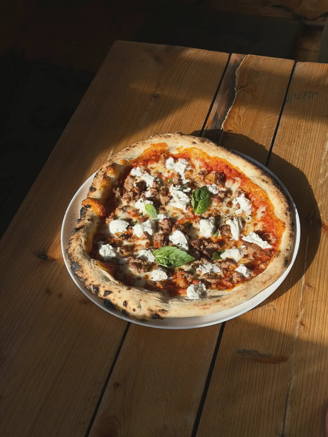 A pizza with crumbled sausage, torn mozzarella and basil on a sunlit wooden table
