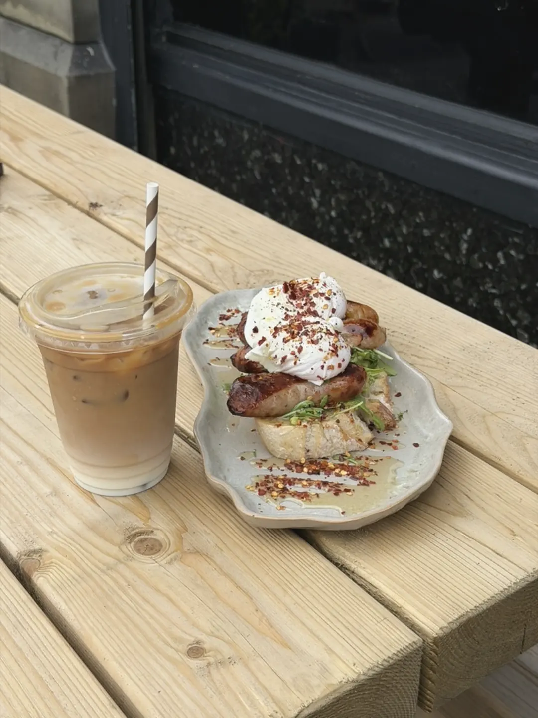 Poached eggs and sausage on toast with chilli flakes, next to an iced latte, on a picnic table outside