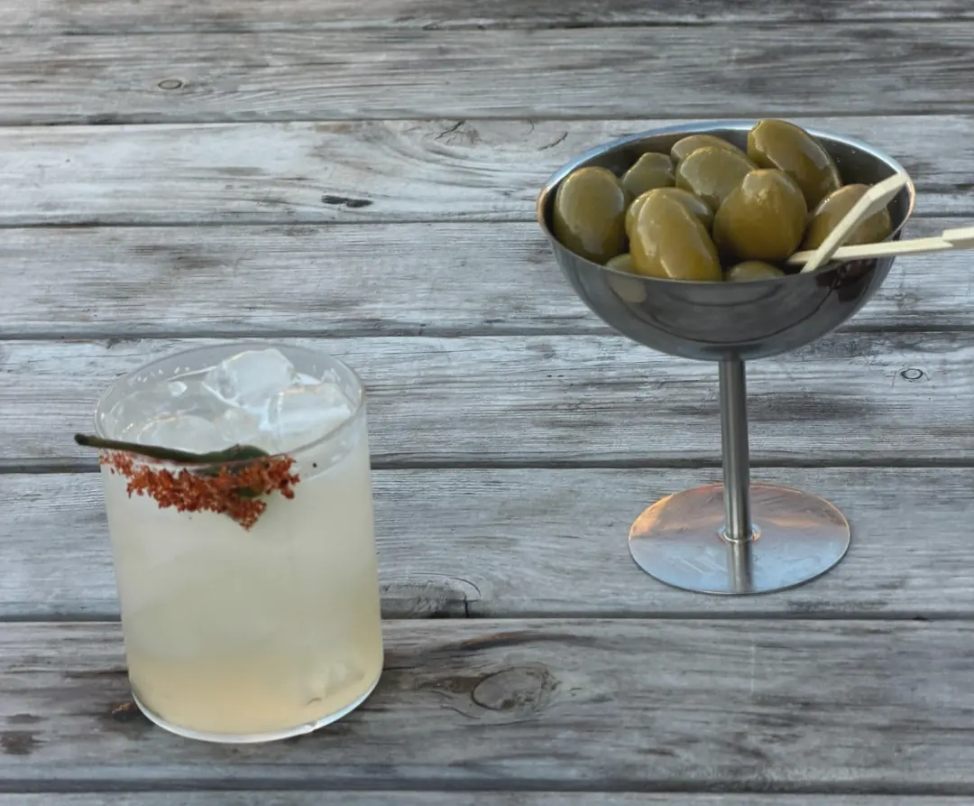 A spicy margarita with a chilli-salt rim and a green chilli, next to a bowl of green olives on a wooden table outside
