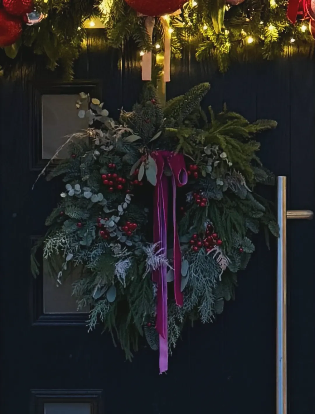 A wreath of pine, cedar and eucalyptus with red berries and a pink velvet ribbon on a dark front door, under a lit garland with red baubles